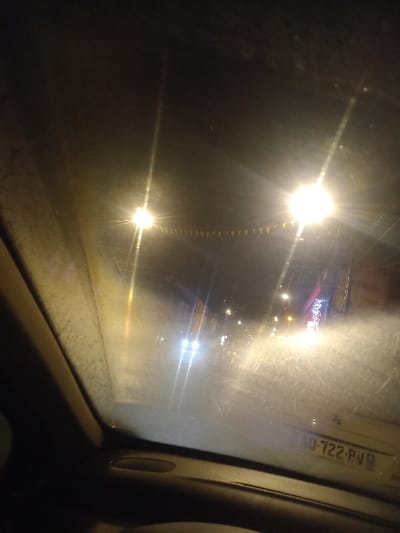 A night view through a car window shows streetlights with starburst effect. Pennants hang between two lights. A number plate is visible on the window's edge. In the distance, blurred lights suggest buildings. The scene has low visibility, possibly due to fog or a dirty windshield. The car's interior is visible along the right edge of the frame.