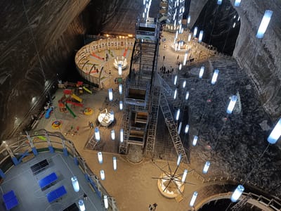 An aerial view shows the Turda Salt Mine in Turda, Romania, an underground entertainment complex within a salt mine. The vast cavernous space is illuminated by numerous hanging cylindrical lights. In the foreground, a court with multiple blue and black ping pong tables is visible. Further back, a playground area with slides and swings is situated on a sandy floor. In the midground, a circular mini-golf course with colorful holes is set up. Dominating the center of the image is a large, industrial-looking Ferris wheel structure, with its cabins visible. Several people are scattered throughout the complex, walking, standing, or engaging in activities. The textured, rough walls of the salt mine rise up on either side, creating a dramatic and unique backdrop to the recreational facilities.