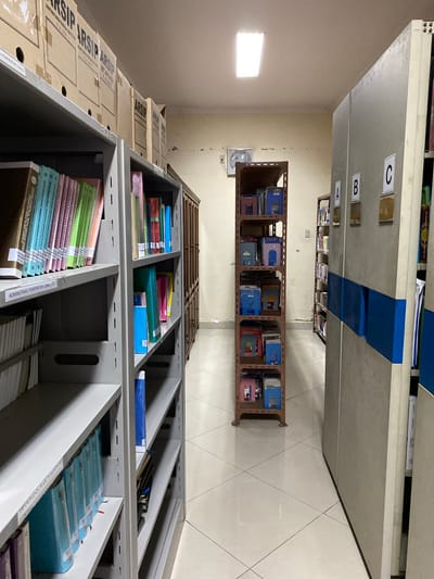 This image shows a dimly lit archive room filled with filing cabinets and shelves, packed with documents and files. The room appears to be a government or institutional archive, as indicated by the labeling of documents and boxes.
In the foreground, large cardboard boxes labeled "ARSIP" (Indonesian for "Archive") are stacked on a metal shelf. Further back, rows of metal shelves hold various binders and files, some with colorful covers, others appearing to be bound reports. Some shelves are organized with folders and individual documents, while others contain what appear to be boxed collections.
The filing cabinets in the background are grey metal units with labels "A," "B," and "C" on their drawers. The presence of labels like "PEMERINTAH LOKAL" (Local Government) and "ADMINISTRASI PEMERINTAH LOKAL" (Local Government Administration) suggest the archive contains records related to local governance in Indonesia. The overall impression is one of organized, if somewhat dense, storage of official records. The lighting suggests an interior scene, possibly during daytime, given the light coming from a window. The room is tiled, indicating an institutional setting. There are no people visible in the image.