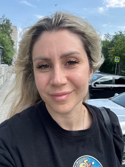 This media file is a portrait of a young woman. She has blonde hair and is wearing a black t-shirt with a graphic on the left side. A thin silver necklace is visible around her neck. She is looking at the camera with a slight smile. The image appears to be taken outdoors during the day, with a cloudy sky and green trees in the background. A yellow pedestrian crossing sign is visible in the upper left portion of the image. There is a parked car to the right of the woman. The location is identified as Novoaltaysk, Russia.