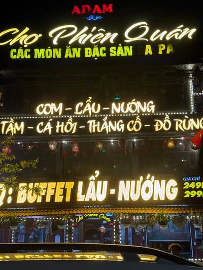 The image shows a brightly lit restaurant sign in Sa Pa Ward, Vietnam. The sign reads "ADAM Chợ Phiên Quán CÁC MÓN ĂN ĐẶC SẢN A PA," followed by "COM - CẦU - NƯỚNG," "TÂM - CÁ HỒI - THẲNG CỐ - ĐỒ RÙNG," and ": BUFFET LẨU - NƯỚNG." The sign advertises a buffet with prices listed as 249 and 299. The restaurant facade is decorated with lights and greenery. Other store signs are also visible.