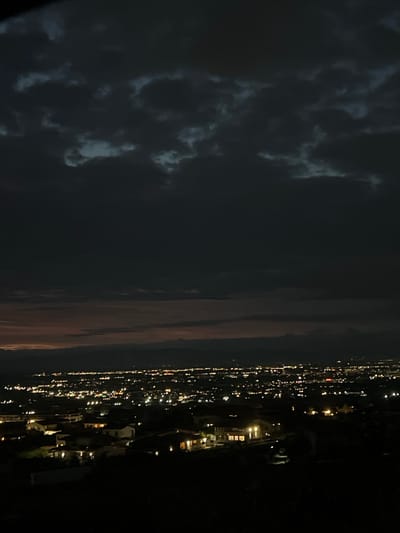 The image is a nighttime aerial view of a city, likely Markovo, Bulgaria, illuminated by numerous artificial lights. Dark, ominous clouds fill the upper portion of the frame, obscuring the sky. In the mid-ground, a dense network of buildings with lit windows creates a sparkling urban landscape. The foreground features a darker, silhouetted area of land with a few more illuminated structures. The overall impression is one of a sprawling city under a cloudy night sky, with the lights of civilization contrasting against the darkness.