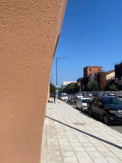 The image captures a street scene in Thessaloniki, Greece, likely during daylight hours given the clear blue sky and strong shadows. The foreground is dominated by a textured, light brown wall, suggesting an architectural element or a garden boundary. Beyond this wall, a row of parked vehicles, primarily cars and possibly some vans or SUVs, line a paved street. The vehicles appear to be modern.

In the background, buildings of varying architectural styles are visible. Some appear older, with brickwork and industrial-like features, while others are more modern and lighter in color. Greenery, in the form of trees, dots the landscape between the buildings and along the street, adding a touch of nature to the urban environment. One of the buildings seems to have an industrial or historical look, possibly an old factory or warehouse. The sky is a vibrant, clear blue, with a faint, almost imperceptible smudge that could be the moon or a cloud, adding a subtle detail to the otherwise cloudless expanse. The overall impression is a sunny day in a city with a mix of old and new architecture, a common sight in many urban areas. There is no visible text in the image.