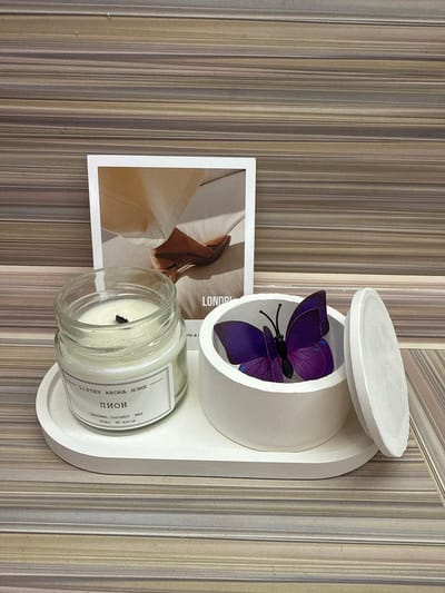 A white oval tray sits on a horizontally striped brown and gray surface. On the tray are a clear glass jar with a white candle inside, a white cylindrical container with an open lid, and a postcard. The candle jar has a label that reads "LISTEN AROMA HOME" and "ПИОН" in Cyrillic. Next to the candle, the white container holds a purple butterfly decoration. The postcard behind the objects features an image of a person's feet in brown shoes and the text "LONDR" and "TO B I" partially visible. The overall scene is a still life arrangement, likely for decorative purposes, placed indoors. The background appears to be a wall with a wood-like striped pattern.