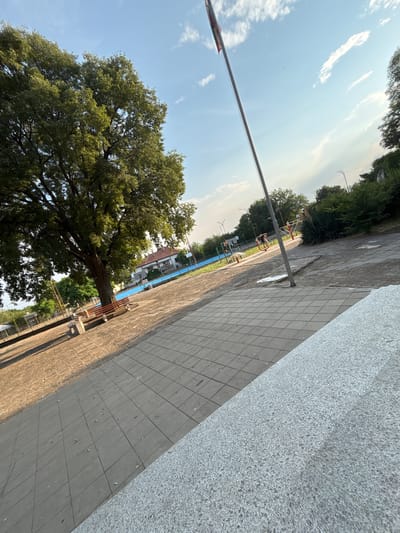 The image captures an outdoor scene in the city of Bulgarovo, Bulgaria, on a sunny day with a clear sky and scattered clouds. The foreground is dominated by a textured gray stone structure, angled from the bottom right corner upwards. To the left of this structure, a paved area with rectangular gray tiles stretches into the mid-ground.
A large, leafy green tree stands prominently in the left mid-ground, casting shadows on the paved area. Adjacent to the tree, a brown wooden bench sits on a concrete base. Further into the mid-ground, a blue rectangular area, possibly a swimming pool or court, is visible. Behind the blue area, a building with a red roof is partially obscured by trees.
In the right mid-ground, a flag pole with a flag is erected. Several pieces of outdoor exercise equipment, painted in red and yellow, are present. Two individuals are engaged in activities on this equipment. More trees and greenery line the background, with a street lamp visible in the distance. The overall environment appears to be a public space, possibly a park or recreation area.