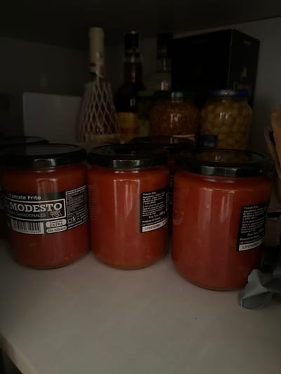 Three jars of "Tomate Frito" from "El Modesto" brand are prominently displayed on a shelf. The jars contain a red, chunky tomato sauce. The labels are visible, with one on the left showing "El Modesto Tomate Frito" and the other two on the right displaying "Tomate frito" and "Producto de España". The jars are arranged from left to right. Behind the jars, other items are visible, including what appears to be other food products in jars and bottles. The shelf appears to be inside a pantry or cupboard. The overall scene is dimly lit. The image does not provide any information or context that would allow for a location in the city of Murcia, Spain.
