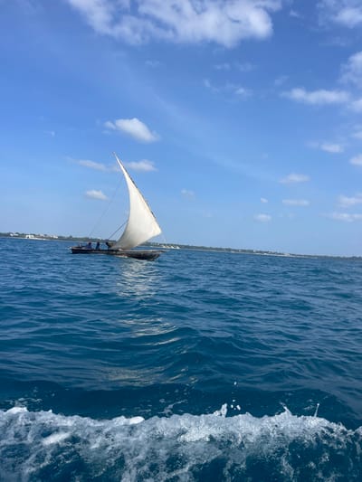 The image depicts a daytime marine scene off the coast of Zanzibar, Tanzania. A traditional wooden sailboat, likely a dhow or ngalawa, with a single white triangular lateen sail fully unfurled, navigates calm, deep blue waters from left to right across the frame's mid-ground. At least three individuals are visible aboard the sailboat; one is positioned centrally near the mast, while two others are further aft. The vessel appears to be propelled by wind, creating a subtle wake. In the immediate foreground, turbulent white foamy water indicates the observation point is from another moving vessel. The distant background reveals a low-lying coastline stretching horizontally, marked by light-colored buildings and sparse green vegetation under a clear blue sky with scattered white cumulus clouds.