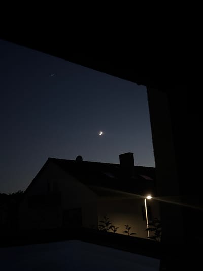 A crescent moon is visible in the dark night sky above a silhouetted house. The house has a pitched roof with visible skylights and a chimney. A single streetlamp illuminates a portion of the house's exterior, casting a bright light and creating lens flare. Some small plants are visible in the foreground, also illuminated by the streetlamp. The overall scene appears to be a residential area in the city of Wörth am Main, Germany. There are no people or animals visible in the image.