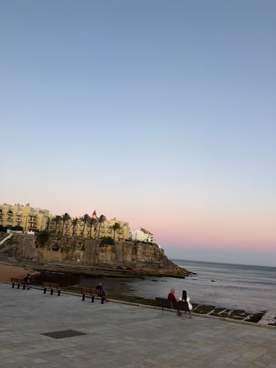 The image shows a scenic view of a coastline with buildings perched on a cliff overlooking the ocean. The sky transitions from a pale blue to a soft pink and orange hue, suggesting either sunrise or sunset. On the left, a row of cream-colored buildings with balconies and palm trees are visible against the rocky cliff face.
In the foreground, a paved walkway runs along the edge of the water. Several benches are placed along this walkway. On one bench, a man in a red shirt and dark pants is seated, facing away from the camera. Another bench is occupied by two figures, one in white, both facing the sea. The water is calm, with a rocky shoreline.
The scene evokes a sense of tranquility and leisure, likely a public space for relaxation and enjoying the coastal views. The time of day appears to be either early morning or late evening, given the sky colors. The location context provided is Cascais, Portugal, which is known for its beautiful coastline. No visible text is present in the image.