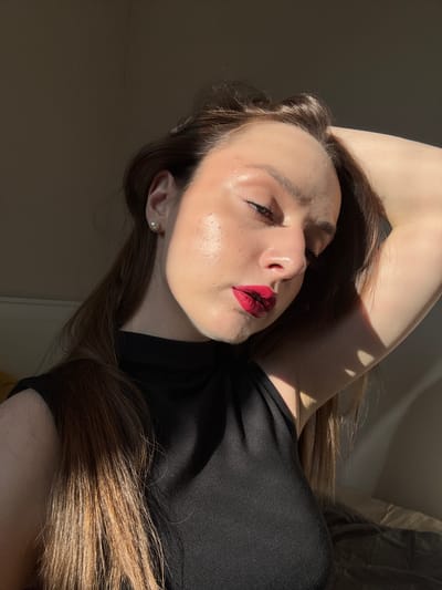 A young woman with long brown hair and fair skin is captured in a sunlit indoor setting. She is wearing a black sleeveless top. Her right arm is raised and bent behind her head, with her hand resting on her hair. Her head is tilted down and to the right, with her eyes closed and her lips painted with bright red lipstick. Sunlight illuminates her face, highlighting her cheekbones and the bridge of her nose. She is wearing a small pearl earring in her visible left ear. The background is a plain, dark wall with a bed visible on the right side, suggesting the location is a bedroom. The image appears to be a close-up, self-portrait. There are no visible elements that would indicate a specific location within Palilula Urban Municipality, Serbia.