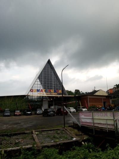 This image, identified by the user as located in Dawathang_Dorjibi_ Kashingtsawa, Bhutan, depicts an outdoor scene featuring a commercial establishment under an overcast sky.

The central subject is a large building with a distinctive triangular, possibly pyramidal, glass facade forming its upper structure, characterized by a visible grid pattern. Below this glass section, a horizontal sign displays the multi-colored text "GUMATI PUNCAK" in prominent letters, with "RESTO KHAS SUNDA" written directly beneath it. The lower section of the building features a wall adorned with green foliage or a vertical garden, and a line of string lights is visible along its length. An entrance area with white architectural elements is positioned beneath the sign.

In front of the building, a dark-paved parking lot contains at least seven parked passenger vehicles of various models and colors, including sedans and a minivan. A tall, dark, curved street lamp post with a modern fixture stands in the mid-ground, slightly to the right of the building's center. To the right side of the main building, a smaller, single-story brown brick building with a reddish roof is visible, with multiple motorcycles parked in its vicinity. Further in the background, additional buildings and dense green trees are present.

The immediate foreground consists of uneven terrain covered with wild green vegetation and some concrete block structures. A partially obscured white banner with faint red text is visible on the far right. The sky is entirely overcast with heavy grey clouds, resulting in dim, diffused lighting across the entire scene. No individuals are clearly discernible, and no specific actions or interactions are depicted.