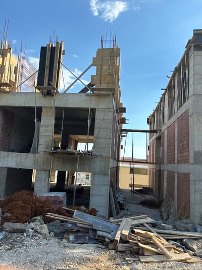 The image shows an outdoor construction site under a clear blue sky with scattered white clouds. The scene depicts a partially built concrete structure, likely an apartment building, with scaffolding and formwork still in place. There are exposed concrete columns, beams, and slabs, with some sections showing brickwork. Piles of construction materials, including lumber, rebar, rocks, and debris, are visible on the ground. The sun appears to be high in the sky, suggesting it is midday. The overall impression is one of an active construction project in progress. There is no visible text in the image.