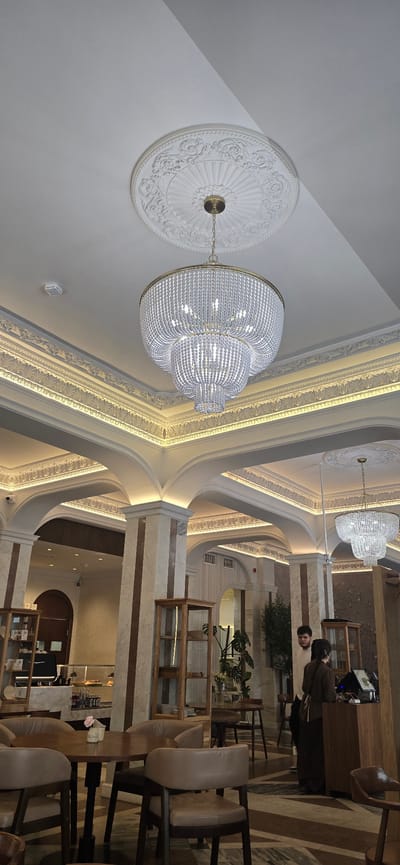 This is a photograph of the interior of a cafe in Pyatigorsk, Russia. The cafe features high ceilings with ornate molding and lighting fixtures. Two large, multi-tiered chandeliers made of clear beads hang from the ceiling. The walls are decorated with marble-patterned pillars and decorative molding. In the foreground, there are several plush, light brown upholstered chairs around round wooden tables. In the background, there is a counter area with a cash register, display cases with merchandise, and a few potted plants. Two individuals are visible in the background, one man and one woman, appearing to be interacting with the counter staff or customers. The overall impression is one of a well-decorated and possibly upscale establishment.
