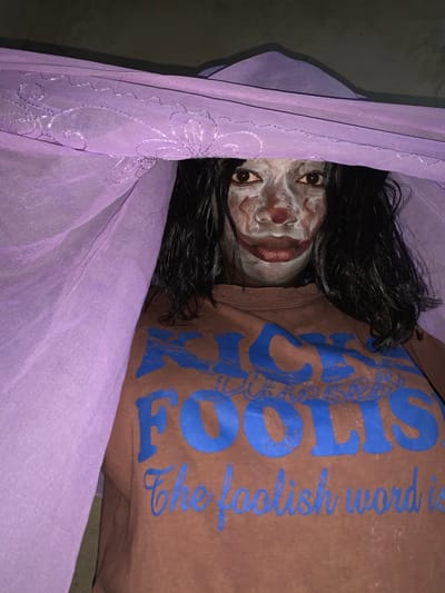 A close-up, partially obscured view of a person with white face paint and dark hair is visible through a purple, sheer fabric, possibly a curtain or mosquito net. The person is wearing a brown shirt with blue lettering that partially reads "FOOLS...the foolish word is". The background is dark, suggesting a low-light environment. Based solely on the provided information, the location and any connection to Agban, Nigeria, cannot be determined.