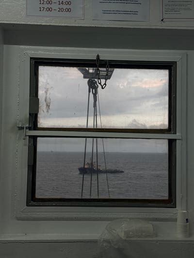 A split-level window looks out onto a body of water. The upper pane of the window shows a cloudy sky and what appears to be a crane structure with chains. The lower pane reveals a boat on the water. The window frame is white and shows some wear and tear. On the windowsill below the window, there are several stacked paper cups and a plastic bag containing more paper cups. A sign with text is visible above the window, showing operating hours in a format of "12:00 – 14:00" and "17:00 – 20:00". Another sign to the right has text related to "VIDEO CALLS PROHIBITED". The overall environment appears to be indoors, possibly on a ship or a building overlooking the sea. The location is specified as Unknown, Italy.