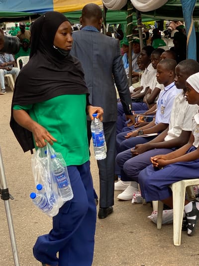 This image captures an outdoor gathering in Shaba, Nigeria, featuring numerous individuals, primarily students and adults, under temporary shelters.
In the foreground, a woman wearing a black hijab, a green long-sleeved top, and blue trousers stands with a plastic bag containing several "Aquafina Pure Drinking Water" bottles in one hand and a single bottle in the other. A black face mask rests under her chin. Her exposed arm shows faint patterned markings, possibly mehndi. Partially behind her, a man in a dark blue suit jacket and light blue shirt is visible from the back. The ground beneath them is paved.
Further back, a group of young people, presumably students, are seated attentively on white plastic chairs. They are dressed in school uniforms, consisting of white shirts (some with blue accents or logos) and blue trousers or skirts. Several students wear white head coverings. Most are looking downwards, suggesting they are listening to a speaker or observing an activity in front of them. One student wears a watch on his left wrist. Another man in a dark suit jacket is seen interacting with the seated students.
The event is set up under tents or awnings, characterized by colorful blue, green, and yellow fabric, indicating an outdoor arrangement. Additional adults are visible in the background, some wearing green shirts or caps, and a large camera lens suggests professional documentation of the event. The scene depicts a structured public gathering, possibly a school assembly, educational program, or community event. The presence of numerous water bottles implies a potentially long duration or warm weather.