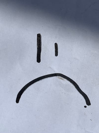 The image displays a close-up of a simple drawing on a textured, light-colored surface, likely paper. The drawing is a stylized sad face, created with thick black marker lines. Two vertical lines represent the eyes, positioned horizontally with a slight gap between them. A single, downward-curving arc forms the mouth, creating the impression of a frown. The lighting suggests the photo was taken indoors, with a gradient of light and shadow across the background, casting a darker tone towards the top. The texture of the surface is visible, showing slight imperfections and speckles. No other objects or individuals are present in the frame. The image does not provide any information that could be used to determine its location in Lanžhot, Czechia, or any other specific city.