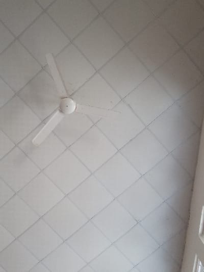 An interior view within the city of Ayama, Nigeria, depicts a white three-bladed ceiling fan centrally positioned in the upper-left quadrant. The fan is mounted on a grid of white square acoustic ceiling tiles, which display dark grout lines or a framing system. Several ceiling tiles exhibit minor dust accumulation or discoloration. A section of an interior wall, featuring multiple window panes, is visible along the right edge of the frame. No individuals or active subjects are present.
