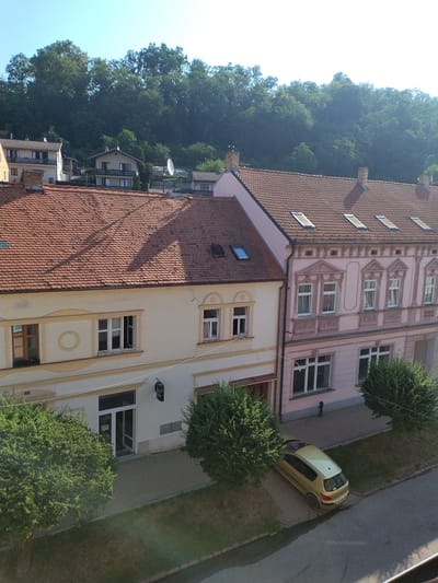 An aerial view captures a street scene in Hluboká nad Vltavou, Czechia, bathed in sunlight. Two buildings line the street; the one on the left is cream-colored with a terracotta tiled roof, featuring multiple windows and a storefront entrance. To its right, a pale pink building with intricate architectural details also has a terracotta tiled roof, punctuated by several skylights. A yellow hatchback car is parked on the cobblestone area beside the buildings. Lush green trees and manicured lawns flank the street and sidewalk. In the background, a dense forest covers a hill under a clear blue sky.