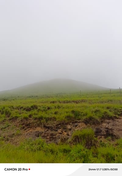 The image depicts a landscape in Nguroje, Nigeria, characterized by a dense fog blanketing a rolling hill covered in lush green grass. A simple wire fence with wooden posts runs horizontally across the mid-ground, separating the foreground from the hill. The foreground features rougher terrain with exposed soil and tufts of grass. The sky is an overcast white, with the fog obscuring any distinct features of the horizon. Text at the bottom of the image reads "CAMON 20 Pro" and "24mm f/1.7 1/207s ISO50".