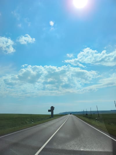 The image shows a view from a car driving on a paved road through a rural landscape. The road has two lanes with white solid and dashed lines. To the left of the road is a green grassy field. On the right side of the road, there are utility poles with wires extending into the distance. In the middle distance, a large, dark, cross-shaped structure is visible on the left side of the road, next to a small signpost. The sky is blue with scattered white clouds. The sun is visible in the upper right corner, causing a lens flare. In the far distance, a dark-colored vehicle is driving away from the camera. The terrain appears to be rolling hills in the background. The scene depicts a sunny day on a highway in a sparsely populated area, possibly in or near the village of Pokrovka, Russia, given the characteristic road markings and rural environment.