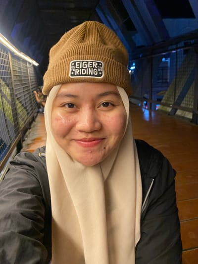 This is a vertical, slightly low-angle, close-up selfie of a young woman wearing a brown beanie and a light-colored hijab. She is also wearing a dark jacket over a lighter garment. The woman has light skin with some blemishes and is smiling faintly. Visible text on her beanie reads "EIGER RIDING EST 1989".
The background is out of focus, showing a dark, indoor environment with some structural elements, possibly part of a building's interior or a covered walkway. There are metal grates and railings visible, along with some artificial lighting that casts a warm glow in some areas and cooler, blue tones in others. The scene suggests an urban setting at night or in a dimly lit area. The overall mood is casual and relaxed.