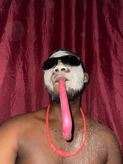 A person lies on a maroon surface, face covered in white powder and obscured by black sunglasses. A pink cylindrical object emerges from their mouth, extending towards the right. An orange beaded necklace encircles their neck and rests on their right shoulder. The person's skin is dark, with white powder also visible on their shoulder and chest. The overall composition suggests an unconventional, possibly artistic, presentation.