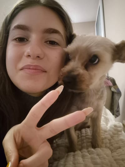 A young woman is taking a selfie with a small, light-brown dog. The woman is holding up her hand in a peace sign, with her fingers near the dog's face. The background is blurry, showing a bed with a textured blanket and a glimpse of a white wall with a monitor in the back. The location is undetermined, and there is no evidence to suggest it is in Pavlikeni, Bulgaria.