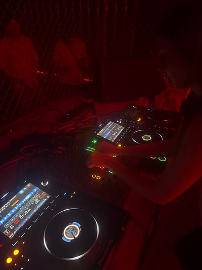 The image displays a dimly red-lit DJ booth within a venue in Shanghai, China. A single individual's forearms and hands are visible, actively operating professional DJ equipment. The setup consists of two Pioneer DJ CDJ media players and a central mixer. The screen of the left CDJ displays a music waveform, the track title "LURGE TO KILL EVERYBODY," a tempo of "175.0," and a playback time of "01:23.388." The right CDJ features an illuminated jog wheel. The DJ's hands are positioned over the mixer's faders and controls, and the left CDJ's interface. On the surface behind the DJ equipment, several clear plastic cups, some containing red liquid, and a white plastic bag partially labeled "Corona" are present. Numerous audio and power cables interconnect the various components. The background features a dark metal mesh or fence structure in the upper left, behind which indistinct, rounded forms are faintly visible under the pervasive red illumination.