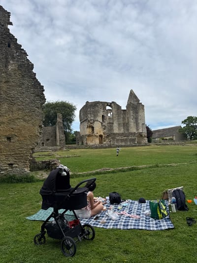 A medium, angled outdoor shot shows a ruined abbey in the city of West Oxfordshire, United Kingdom. In the foreground, a person lies on a blue-and-white plaid blanket with a baby stroller nearby. The abbey ruins are constructed of stone and consist of a tall tower with arched windows, a smaller tower to the right, and a lower stone wall to the left. The sky is overcast with white clouds.