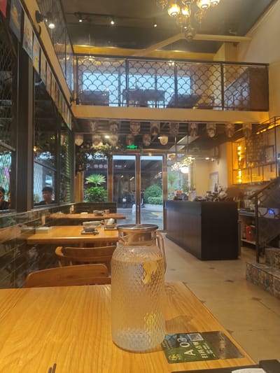 The image depicts the interior of a restaurant or cafe in Nanhai District, China. The foreground features a wooden table with a glass pitcher of water and some coasters. In the midground, several wooden tables are set with small bowls and cups, suggesting a dining or seating area. Further back, a counter or bar area is visible, with shelves and lighting fixtures. The background shows a mezzanine level with seating and a decorative railing. The overall environment appears to be modern and stylish, with warm lighting. There are no prominent actions or interactions captured, and the focus is on the ambiance and decor of the establishment.