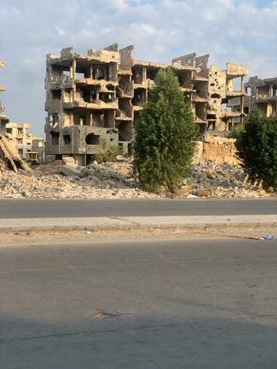 A ruined building stands beside a road. The structure is severely damaged, with many floors exposed and crumbling. Trees grow amid the rubble. Debris litters the ground next to the grey asphalt road. The scene conveys devastation and destruction, suggesting conflict or natural disaster. Sky visible in the background with light cloud cover.