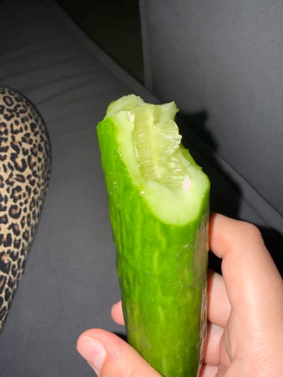 A close-up, waist-up shot shows a person holding a partially eaten cucumber. The cucumber is bright green and has a fresh, moist appearance, with visible seeds and pulp on the bitten end. The person's right hand, with clean fingernails, is holding the lower portion of the cucumber. The person is wearing a garment with a leopard print pattern on their left side, suggesting it's clothing. The background is a dark, indistinct surface, possibly a couch or chair, with a soft shadow cast by the cucumber. The image is well-lit, focusing on the texture and detail of the cucumber and the hand. The scene takes place indoors, with no discernible urban or outdoor elements visible. The location in Kommunarka, Russia is not apparent from the visual information provided in the image.