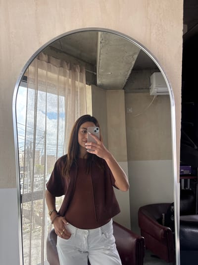 This mirror selfie captures a woman standing in an interior space, likely a salon or a modern room, in Zovuni, Armenia. The image is taken through a large, oval-shaped mirror with a thin silver frame.

The woman, positioned on the right side of the reflection, holds a light-colored iPhone to take the photo, partially obscuring her face. She has long, dark brown hair and is dressed in a dark brown, short-sleeved top or jacket, paired with white pants or jeans. Notable accessories include a watch and bracelet on her left wrist, and rings on her right hand. Her fingernails are painted white.

The interior reflected in the mirror features textured, light beige or grey walls. A white air conditioner unit is mounted high on one wall, with an electrical wire visible beneath it. To the right, part of a dark brown, upholstered chair is seen, suggesting a seating area. Further in the background, a sheer, light-colored curtain or blind covers a window. Through the window, a daytime outdoor scene is visible, including trees, distant buildings, and a clear sky. A small purple box is visible in the upper right corner of the reflection. The overall lighting suggests it is daytime. No discernible text is visible within the image.