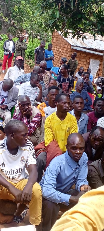 A group of men are gathered outdoors, possibly at a meeting or community event. Most are seated or squatting on the ground, dressed in casual clothing, including t-shirts, button-down shirts, and trousers. Some men stand behind the seated group, including at least two individuals wearing military-style uniforms. A modest brick building with a tin roof is visible in the background, partially obscured by trees. The overall atmosphere appears serious and attentive.