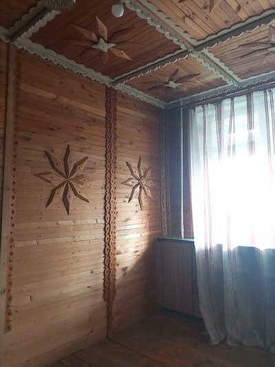 The image depicts the interior of a room, likely a dwelling, with walls and ceiling constructed from natural wood planks. Decorative wooden elements, shaped like stars, are affixed to both the walls and ceiling. The ceiling also features scalloped white trim. A window with light-colored curtains, partially obscuring the view, is visible on the right side of the frame. A wooden cabinet or radiator cover is positioned beneath the window. The floor is also made of wood. The overall aesthetic suggests a rustic or traditional style. The location is Vsevolozhsk, Russia.