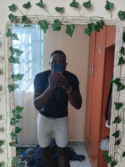 A person is reflected in a mirror decorated with rope and faux ivy. The individual is taking a photo with a blue phone. They wear a black shirt and light-colored shorts. The reflection shows a bedroom with a light brown dresser in the background. Below the mirror, a window is partially visible. A bed and shoes are seen in the lower right corner. The room has neutral-colored walls.