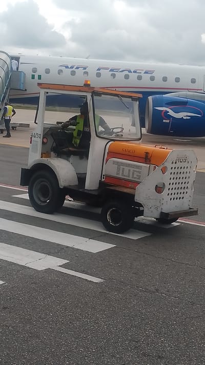 An Air Peace airplane is parked at the tarmac with a tug vehicle in the foreground. The tug vehicle is white with orange accents and a black cab. A person wearing a bright yellow vest and dark pants is seated in the driver's seat of the tug. The tug is positioned on a crosswalk painted with white stripes on the asphalt. The airplane has "AIR PEACE" written in blue lettering on its side, and a blue engine cowling with a white bird logo. The sky is overcast. The location is Orerokpe, Nigeria.