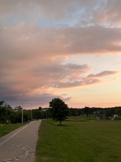 A paved walkway curves through a grassy park with a tree in the foreground and distant trees and buildings. The sky is filled with soft, colorful clouds in hues of pink, orange, and blue. Two people walk on the path in the middle ground, and several streetlights line the walkway. A small body of water with a bridge and some boats is visible in the distance. The scene appears to be set in a park in Carnikava, Latvia, during a sunset.