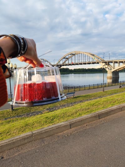 In Bol'shie Mkhi, Russia, a hand with bright red nail polish and multiple bangles on its wrist holds a clear plastic container with a red cake inside, topped with white frosting swirls. The container is secured with white ribbon. The cake is positioned in the foreground, with a river and an arched bridge in the background. A paved walkway, a grassy embankment, and streetlights are also visible. The sky is partly cloudy.