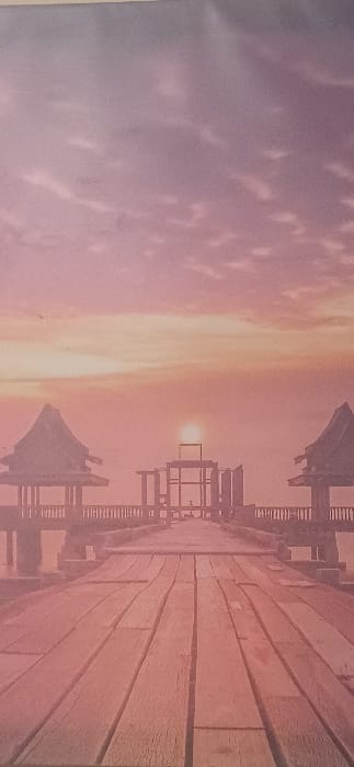 The image depicts a wide-angle view of a waterfront scene at either sunrise or sunset, characterized by dominant pink, orange, and purplish hues. A wooden pier, composed of reddish-brown planks, extends from the foreground towards the horizon. The pier is bordered on both sides by a railing. In the midground, two symmetrical pavilion-style structures with multi-tiered, upward-curving roofs stand as dark silhouettes on either side of the pier. Centered on the horizon, an open, square-framed architectural structure is visible, with the sun, a distinct orange orb, positioned within its upper portion. The sky transitions from a purplish-grey at the top to a bright orange-pink near the horizon, with scattered, wispy clouds. The structures are situated over a calm body of water that reflects the sky's colors. No people, animals, or specific actions are discernible. The architectural style and prominent body of water in the image are not consistent with the known geography or typical structures of Rakevo, Bulgaria, which is an inland village.