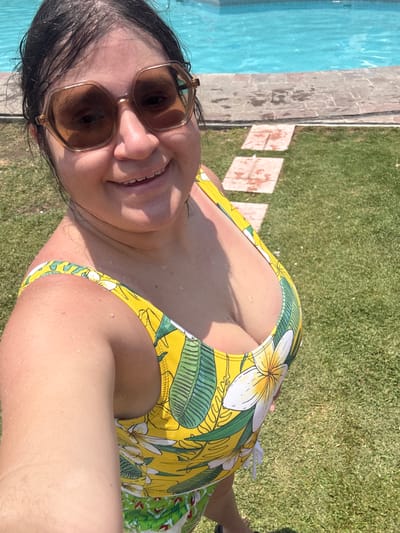 A woman captures a selfie outdoors on a bright, sunny day, likely enjoying leisure time by a swimming pool in Tres de Mayo, Mexico. She is positioned in the foreground, smiling broadly at the camera. Her dark hair appears slightly wet, and water droplets are visible on her skin and shoulders, suggesting she has recently been in the pool or splashed. She wears stylish brown-tinted sunglasses with angular frames.
Her attire is a vibrant yellow swimsuit adorned with a tropical pattern of large green leaves and white frangipani-like flowers. The swimsuit features a plunging neckline.
The background reveals a clear blue swimming pool to her left, with a paved or stone path separating the pool from a patch of lush green grass. The path shows some darker, possibly wet, spots. The overall scene suggests a warm, clear day, with bright sunlight creating visible shadows. No text is visible in the image.