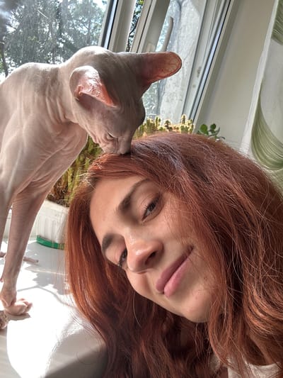 A hairless cat, likely a Sphynx, is leaning its head onto the forehead of a young woman with reddish-brown hair. The woman is looking slightly down and smiling. They are positioned in front of a window with sunlight streaming in. The background includes greenery, possibly plants and trees visible through the window, and a sheer curtain. The scene appears to be indoors, near a window. There is no information or visual cues in the image that would allow for the determination of a specific city or location such as Togliatti, Russia.