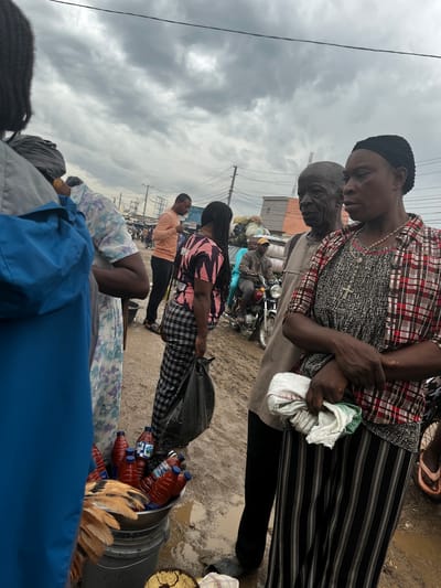 The image depicts an outdoor market scene in Sabojibi, Nigeria, under a cloudy sky. Several individuals, including men and women, are present. One woman in the foreground wears a black skullcap and a red and white checkered shirt, holding a white cloth. Beside her, an older man with dark skin is visible. Further back, another woman is dressed in a pink and black striped top and a checkered skirt, carrying a black bag.

Objects in the scene include several plastic bottles filled with a red liquid, possibly juice or soda, placed in a metal bucket. A motorcycle is parked behind some of the individuals. Bags of produce, including what appears to be yams, are also visible. The ground is wet and muddy, suggesting recent rain.

The overall atmosphere appears to be that of a bustling market. The time of day is unclear due to the overcast weather. The presence of a market, people gathered, and goods for sale are key indicators of the activity. There is no discernible text in the image.