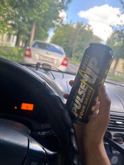 A person's hand holds a black and gold can of "Pulse Up" energy drink inside a car. The can is partially obscured by the steering wheel and the driver's hand. In the background, a white car is visible through the windshield, and trees line a road. The sky is bright with clouds. The dashboard of the car is visible, showing an orange display. The image appears to be taken during the day. The location of Шебунино, Russia, is not determinable from the image content.