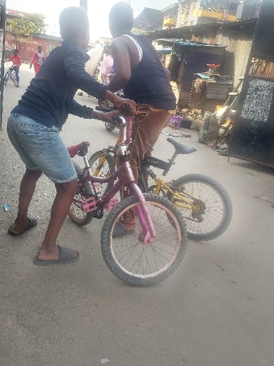 This outdoor photograph, taken in Agani, Nigeria, captures two young males interacting with bicycles on a paved surface during bright daylight.
One boy, wearing a dark t-shirt and light blue denim shorts, stands upright, holding the handlebars of a pink bicycle. He is wearing a black slide sandal on one foot while the other foot appears bare. Next to him, another boy, dressed in a dark t-shirt and brown trousers, is hunched over the front section of the same pink bicycle, appearing to inspect or work on its front wheel or handlebars. The pink bicycle is upright, suggesting it is either being held or momentarily balanced. In the background, a yellow bicycle lies on its side on the ground.
The setting appears to be a street or open area, with a row of informal structures, possibly small shops or dwellings, constructed from various materials including corrugated metal sheets. This suggests a bustling, possibly urban or semi-urban environment. The bright lighting indicates it is daytime with clear weather conditions. Both individuals seem focused on their task. No discernible text is visible within the image.