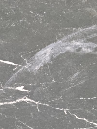 The image shows a close-up view of a dark gray, textured surface with white veins or scratches. The surface appears to be natural stone, possibly marble or granite, with a matte finish. There are several linear white markings across the surface, some thin and sharp, others broader and smudged. In the center of the image, a prominent white streak curves from the upper right to the lower left, appearing as if it was drawn or dragged across the surface. The overall impression is of a weathered or worn material. The image does not provide any information about a specific location in Ivano-Frankivsk, Ukraine, nor does it depict any people, actions, or interactions.