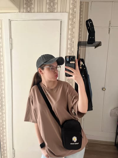 This is a vertical, medium shot of a young person, likely female, taking a selfie in a room with white paneled doors and patterned wallpaper. The person is wearing a light brown t-shirt, a gray baseball cap, and glasses. They have a black crossbody bag with a white daisy design on it. The person is holding a smartphone, which is displaying a pattern of circles and stickers. In the background, a black belt is hanging on a metal rack. The scene appears to be indoors, with natural lighting. There is no indication of the time of day or weather. The person's expression is neutral, and they are looking into the mirror. The text on the bag reads "fashion trends" in reverse.