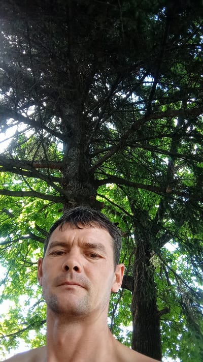 A close-up, low-angle, full-frame, eye-level outdoor shot captures a man's face as he looks directly into the camera. He has short, dark hair with some graying, dark eyebrows, and a weathered complexion. His mouth is closed in a neutral expression. The man is shirtless, with his shoulders and upper chest visible. Behind him, a large, dark tree with thick branches and dark green foliage dominates the background, obscuring much of the sky. The lighting suggests it is daytime, with sunlight filtering through the leaves. The image appears to have been taken with a mobile phone, indicated by the upward angle and the proximity of the subject. The scene does not provide enough information to determine the specific location within Altai, Kazakhstan.