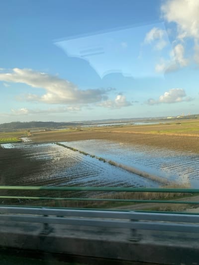 The image displays a panoramic view of extensive, partially inundated agricultural fields within the Montemor-o-Velho region of Portugal, characteristic of the Lower Mondego Valley's floodplains. The vantage point is from an elevated structure, indicated by a green metal railing and a grey concrete barrier occupying the lower foreground. The landscape is composed of dark, tilled earth organized in linear rows, interspersed with substantial sections of shallow standing water reflecting the blue sky and scattered white clouds. In the far distance, low, vegetated hills are visible on the left horizon, while two prominent, slender industrial structures, resembling chimneys or towers, rise against the sky on the right. No individuals, vehicles, or specific activities are discernible within the external scene. The upper portion of the photograph exhibits distinct reflections on a transparent surface, likely a vehicle window, revealing distorted textual elements including "RESERVADO A PESSOAS" and "DE ÁGUA".